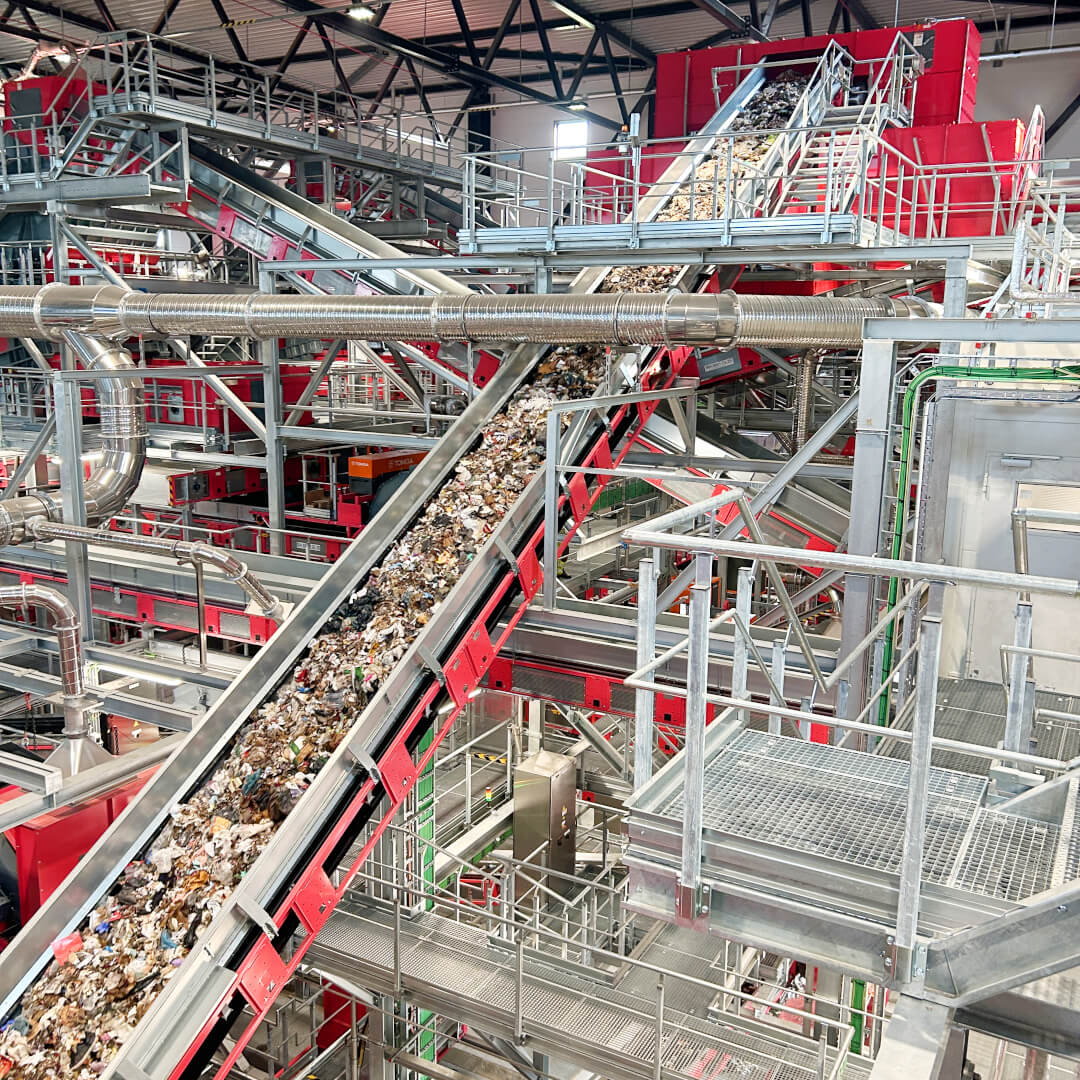 Part of a municipal solid waste sorting plant, shown in red. In the foreground, a conveyor belt transports household waste.