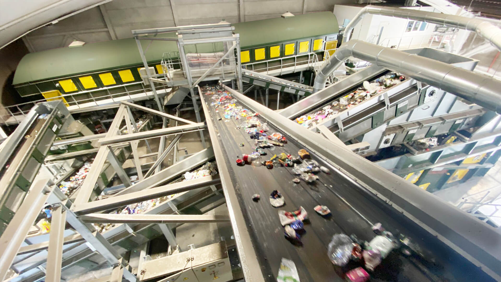 Municipal solid waste (MSW) sorting facility in green and yellow. A conveyor belt carrying household waste is shown in the foreground, while a trommel screen in the background separates the waste into different fractions.